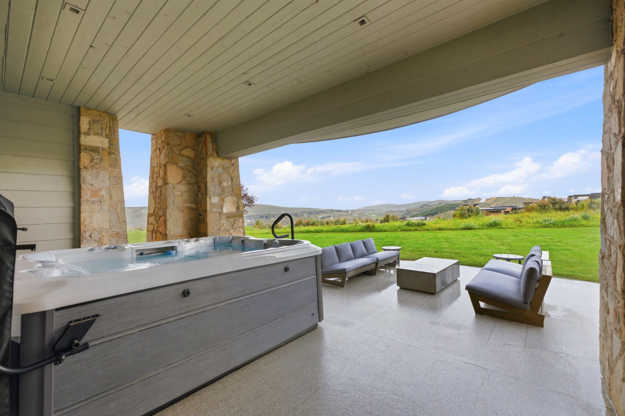 Covered patio featuring a built-in swim spa, stone pillars, concrete flooring, and a vast green landscape