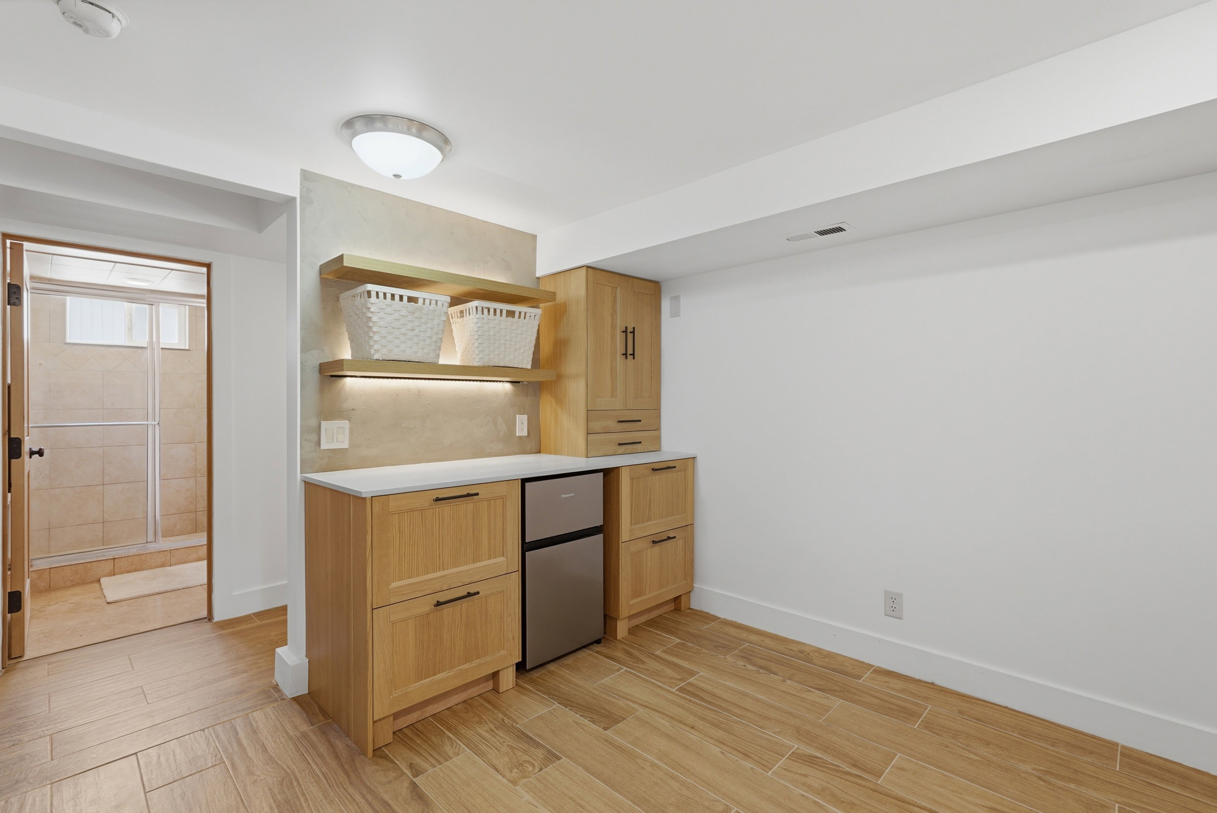 Built-in cabinetry with a light wood finish, white countertop, wet bar with open shelving with integrated lighting, and a compact stainless steel refrigerator
