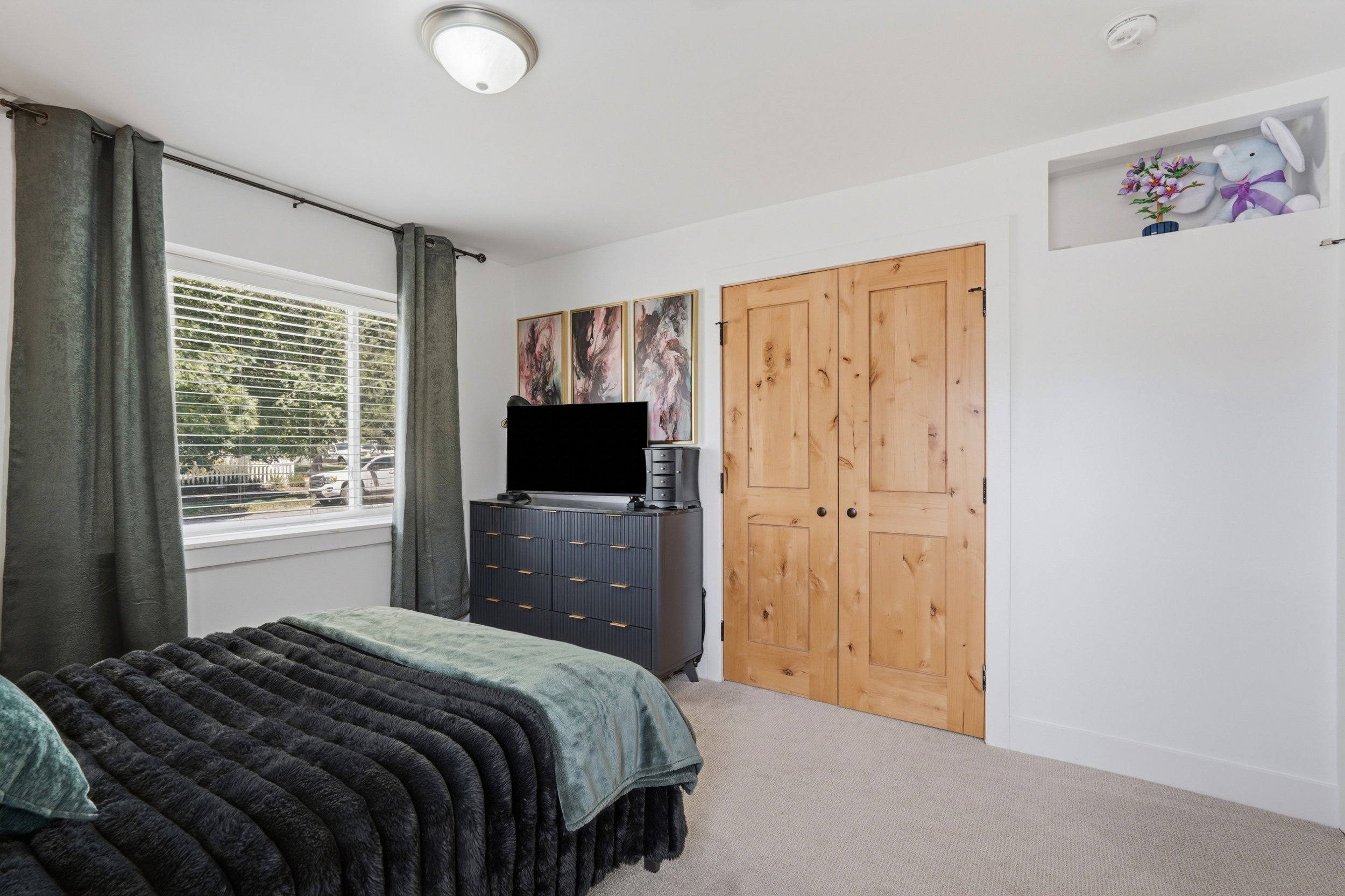 Carpeted room featuring a large window with blinds, natural wood double closet doors, a built-in display nook, and a flush-mount ceiling light fixture