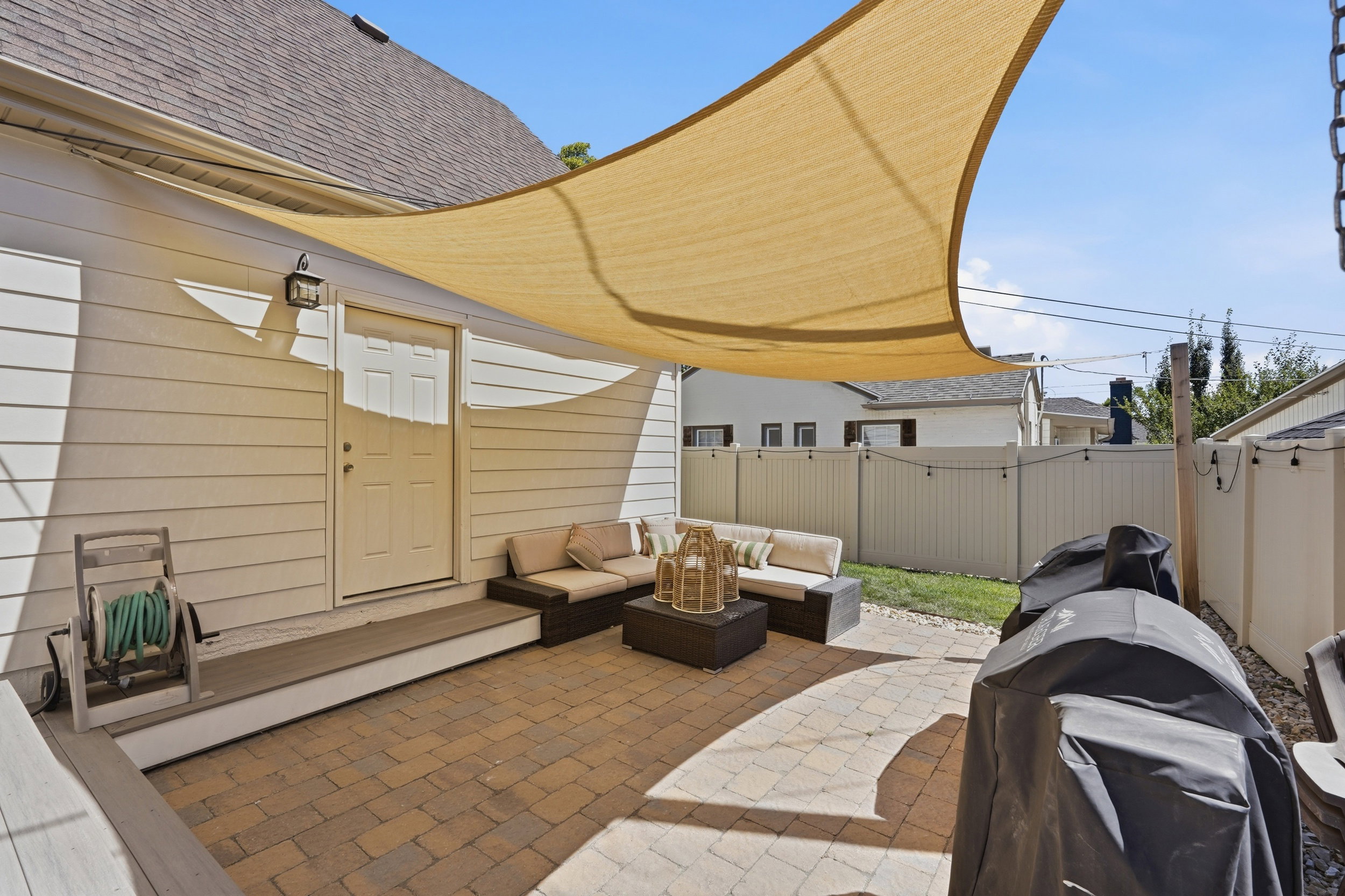 Expansive paver patio with a large sail shade, featuring a raised deck landing, exterior door access, and a durable vinyl fence
