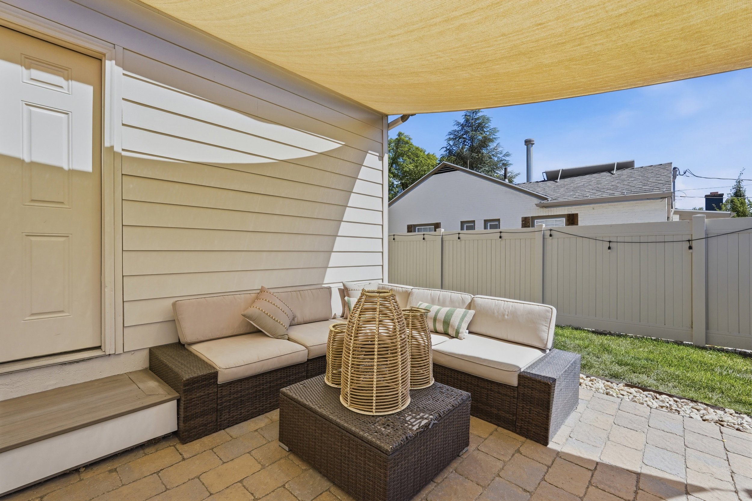 Paved patio featuring a shade sail, tan siding, and a white privacy fence bordering a manicured lawn