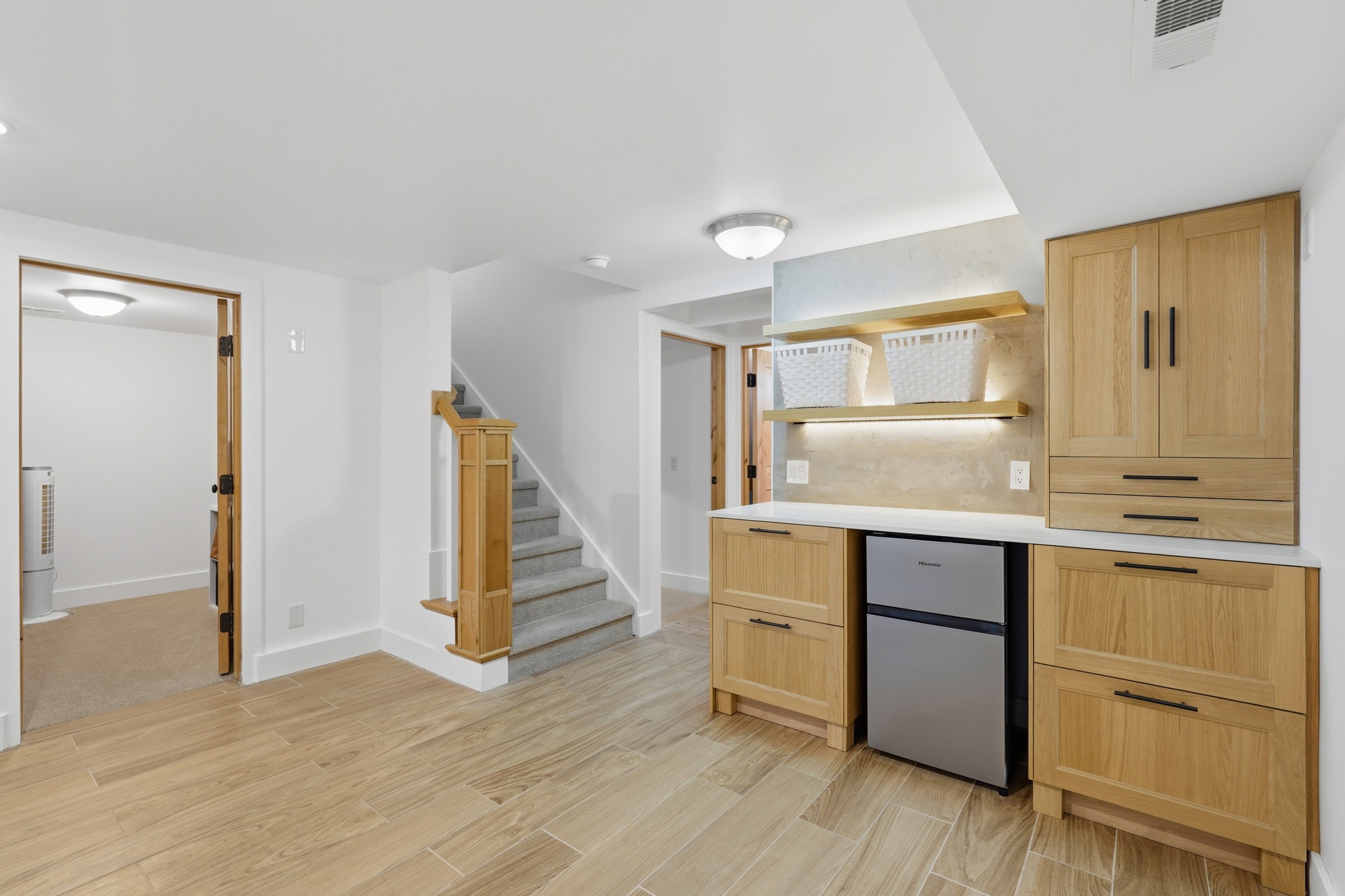 Welcome to the basement space. Built-in cabinetry with light wood finish and dark hardware, featuring a wet bar, with compact refrigerator, open shelving with integrated lighting, and a white countertop