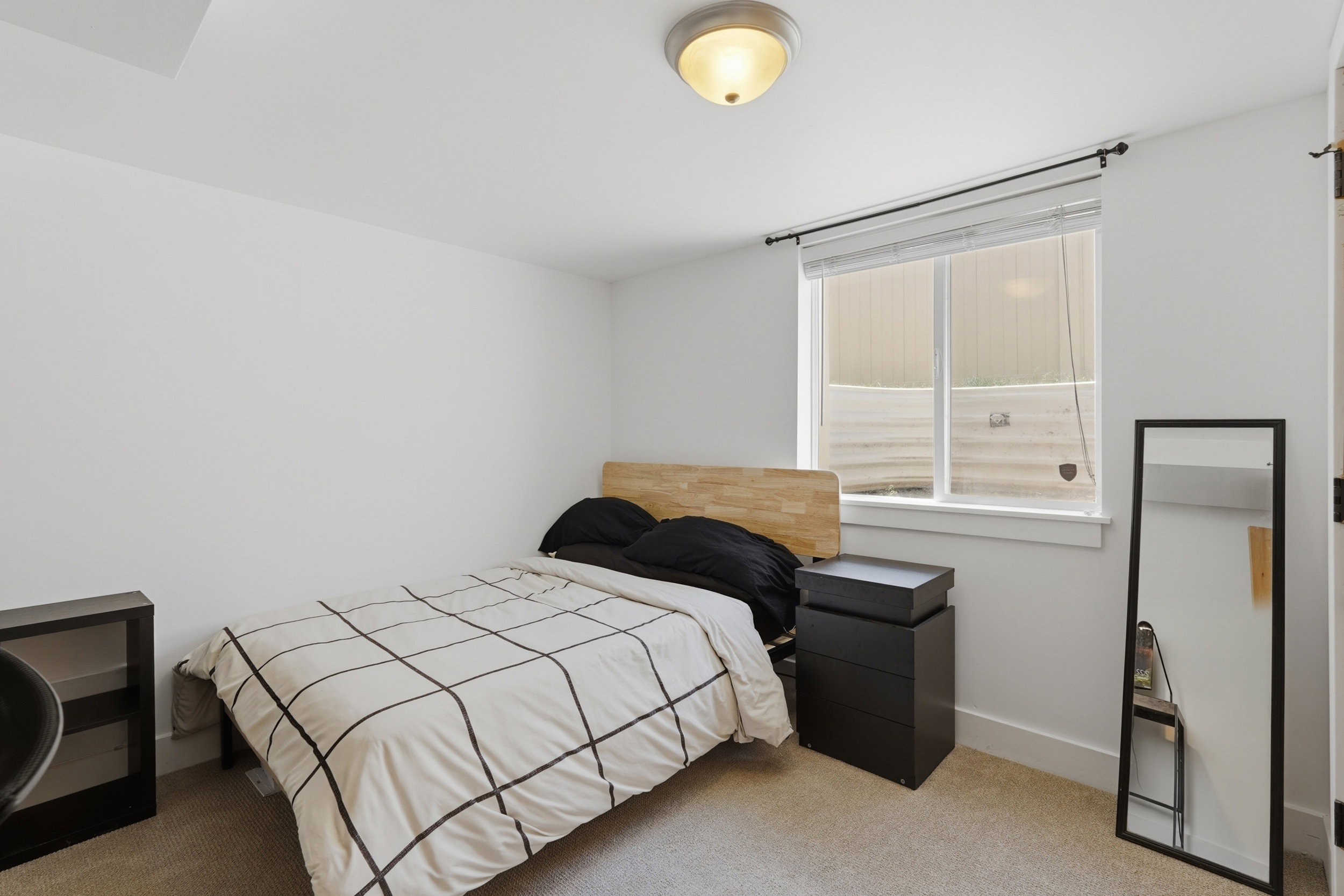 Carpeted room featuring a white ceiling and walls, a flush-mount ceiling light fixture, a window with roller blinds, and a wood-finish headboard