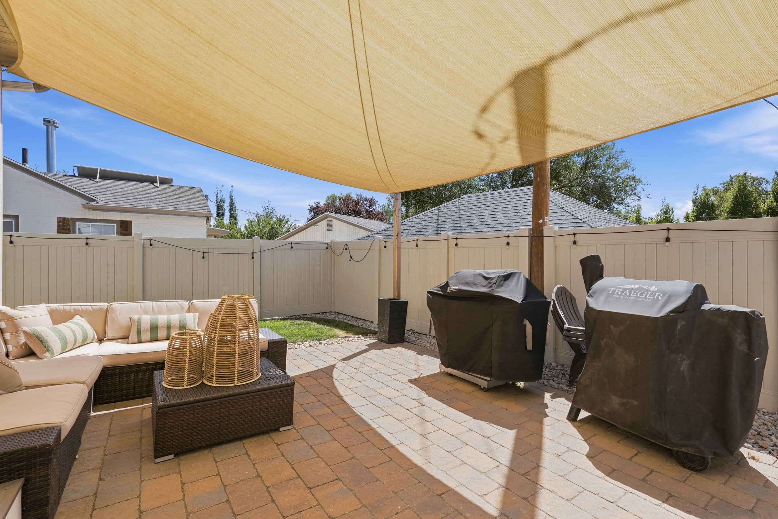 Paved patio area featuring a light-colored shade sail, string lighting, and a privacy fence