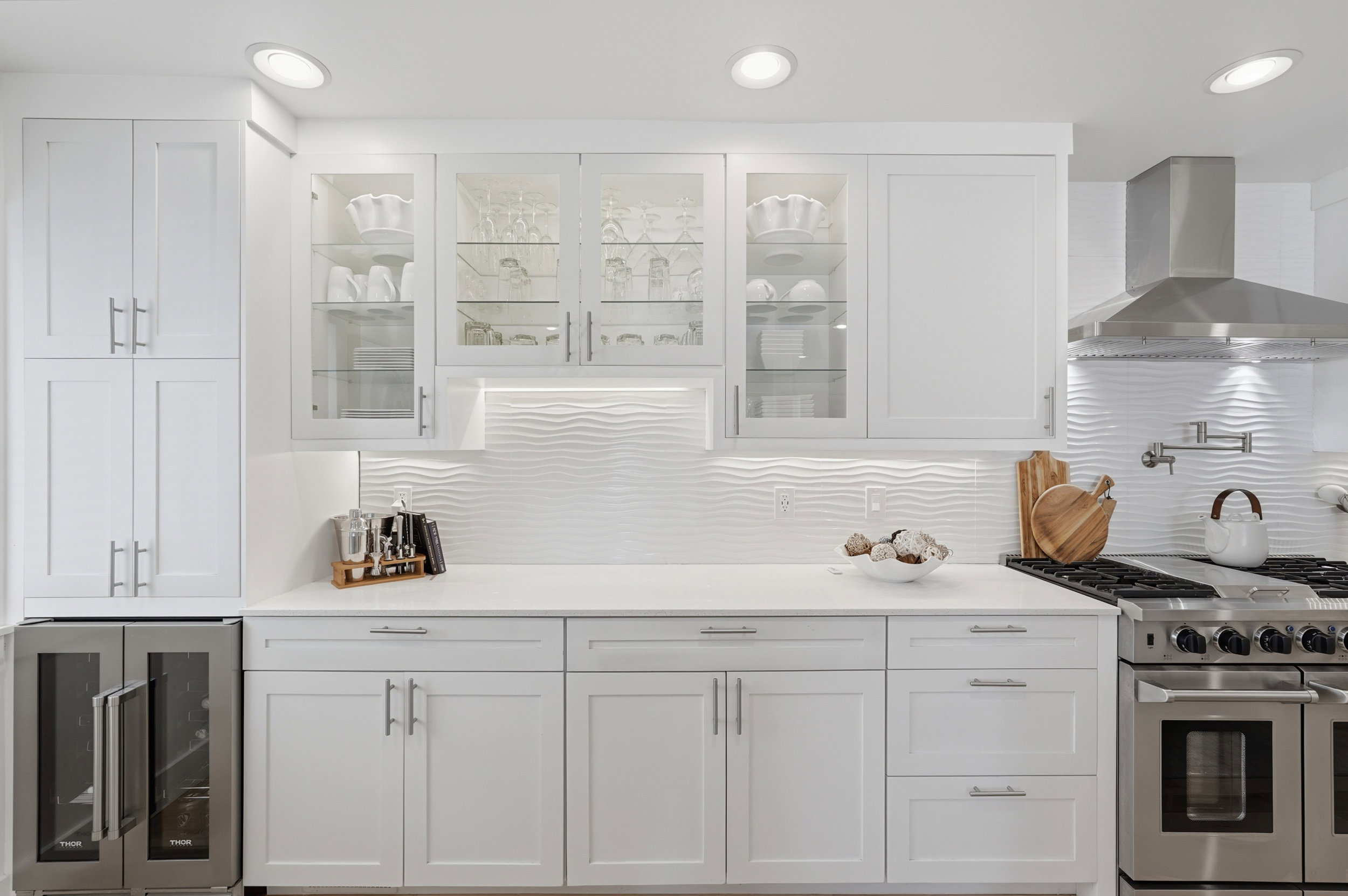 White shaker cabinetry with glass upper doors, stainless steel range with pot filler, complementary range hood, and textured white tile backsplash
