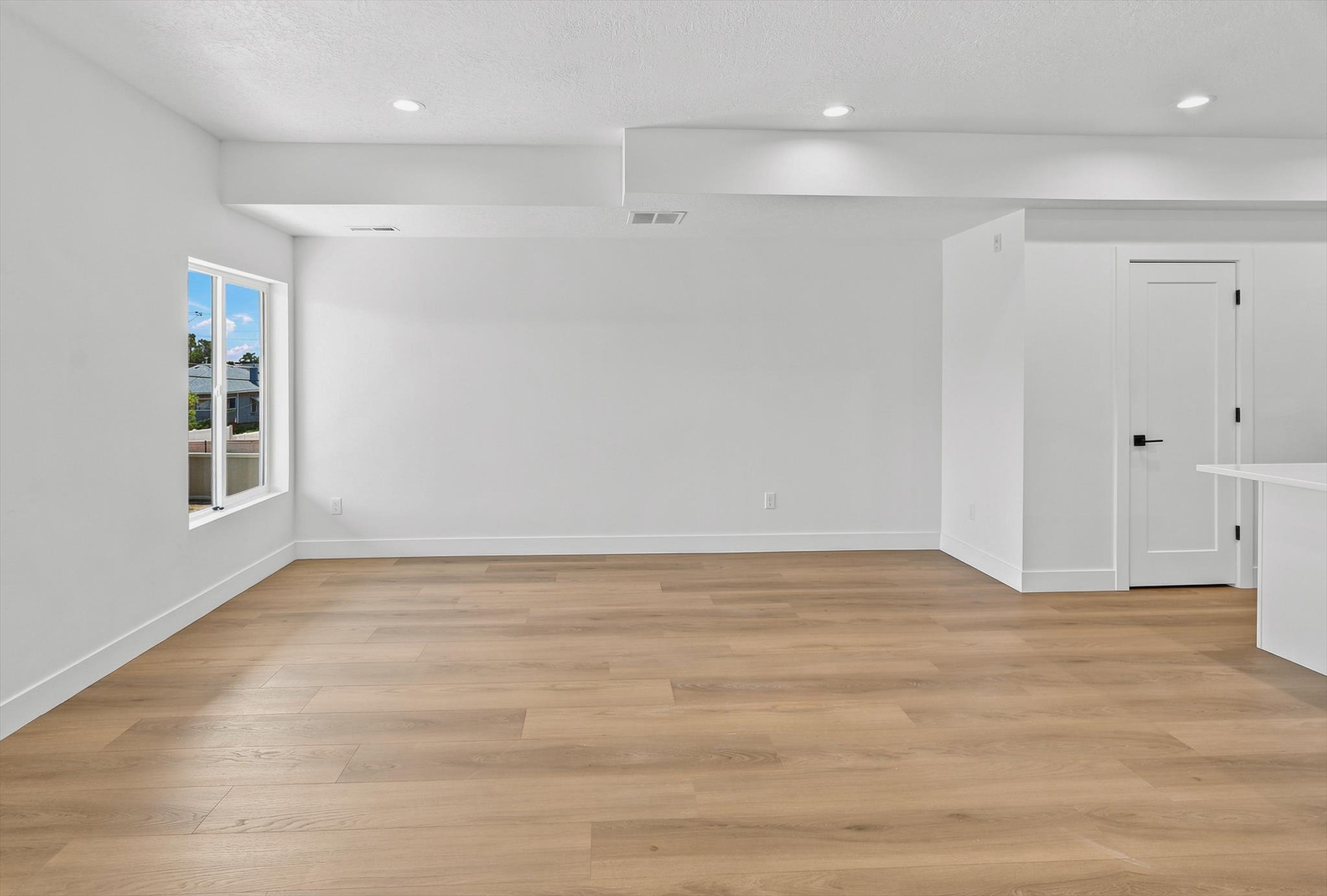 Open-concept room featuring wood-finish flooring, recessed lighting, a white interior door with matte black hardware, and large windows providing natural light