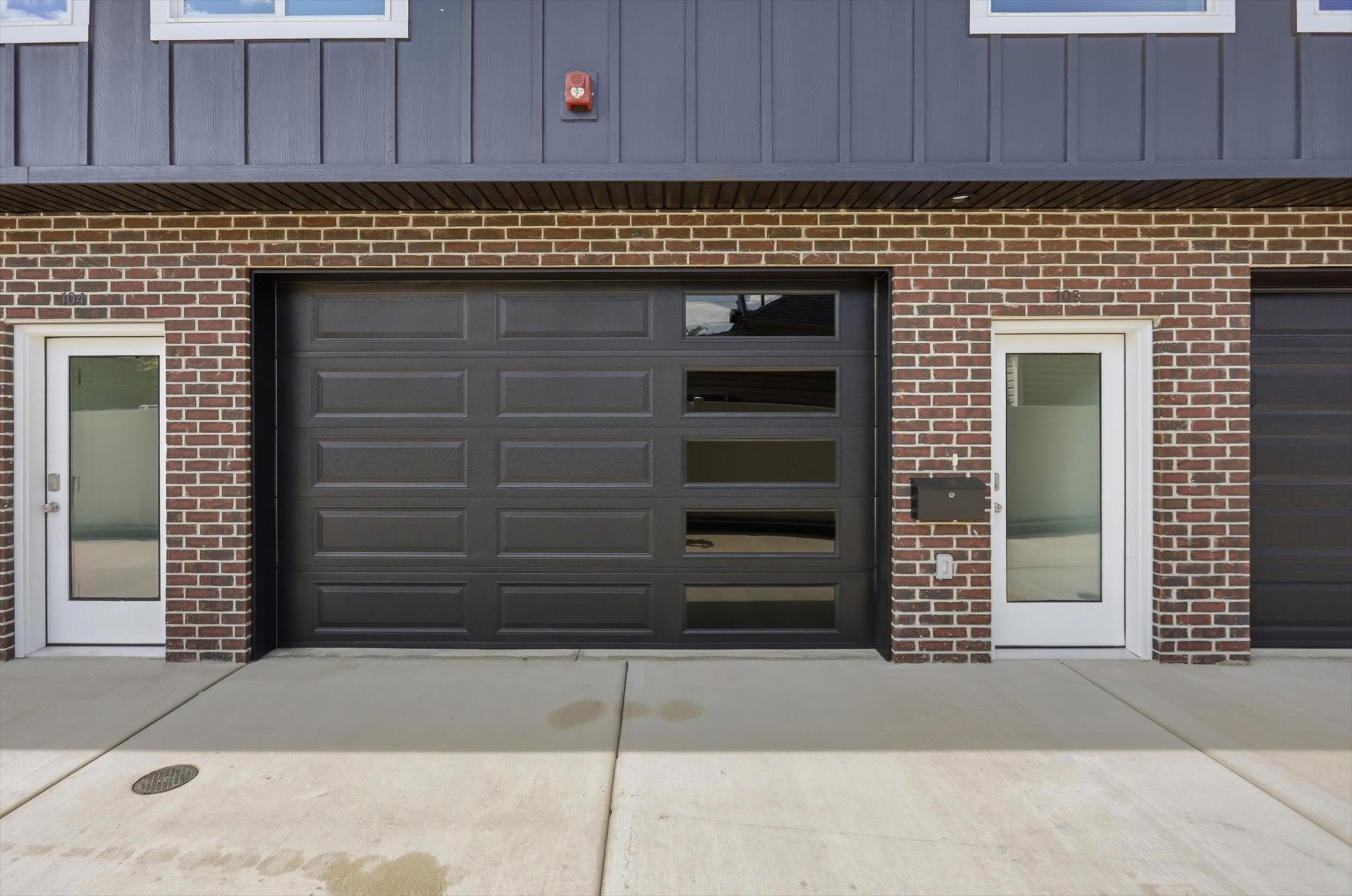 Brick facade with dark vertical siding featuring a modern garage door with rectangular window inserts, a full-lite glass entry door, and concrete driveway