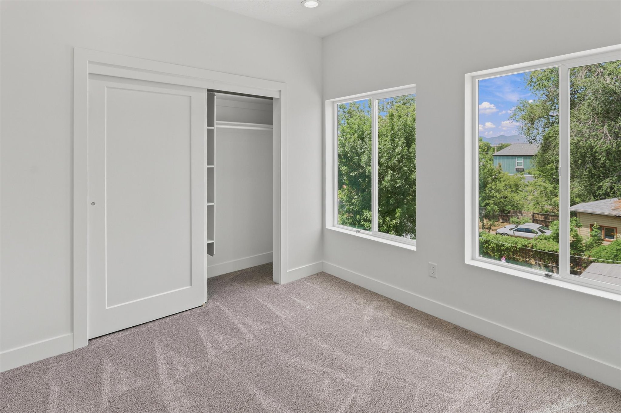 Bright interior space featuring a sliding closet door with built-in shelving, plush gray carpeting, white baseboards, and two large windows offering extensive outdoor scenery
