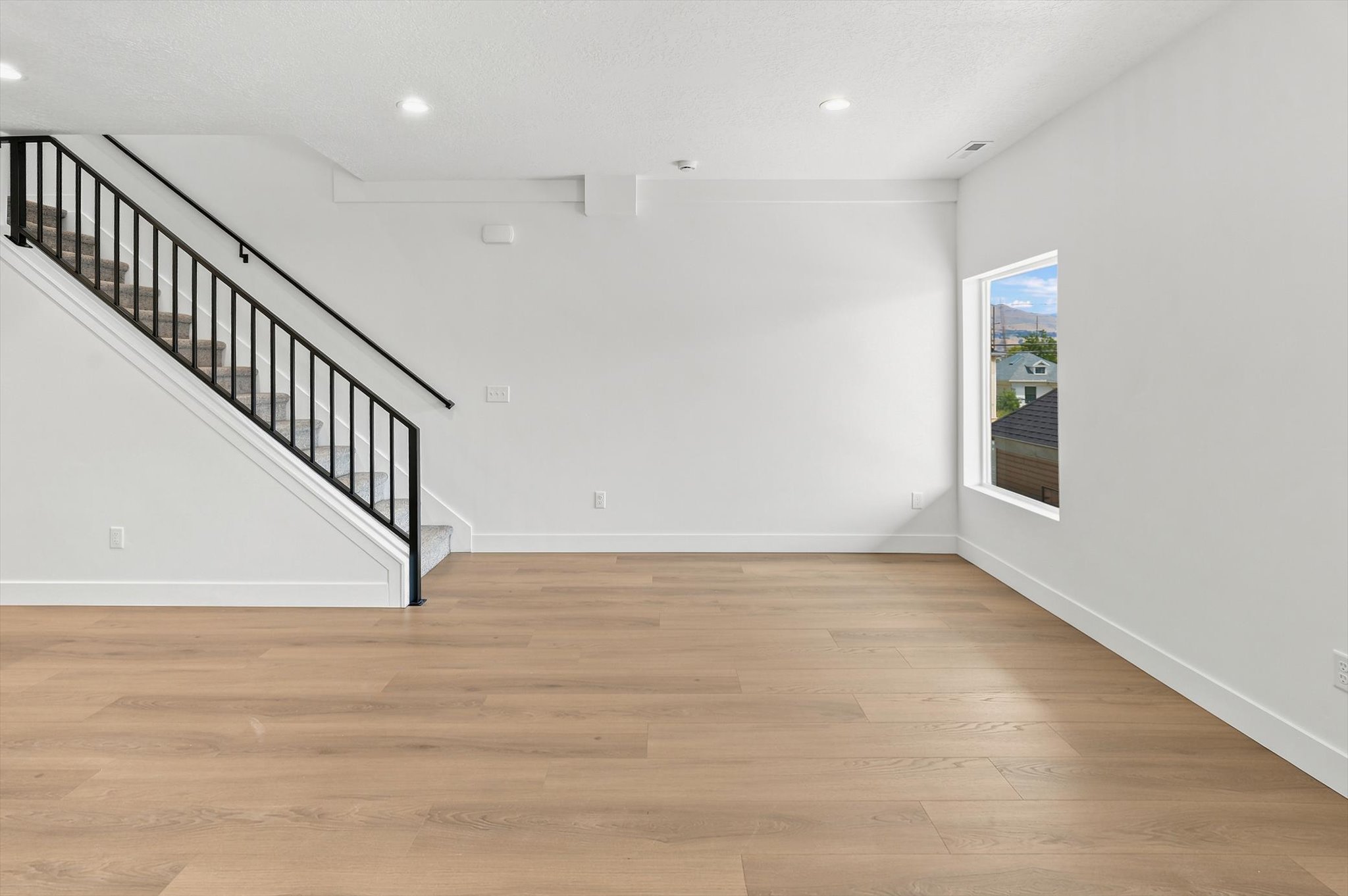 Spacious room featuring light wood-finish flooring, a contemporary staircase with black wrought iron spindles, abundant natural light from a large window, recessed lighting, and crisp white baseboards
