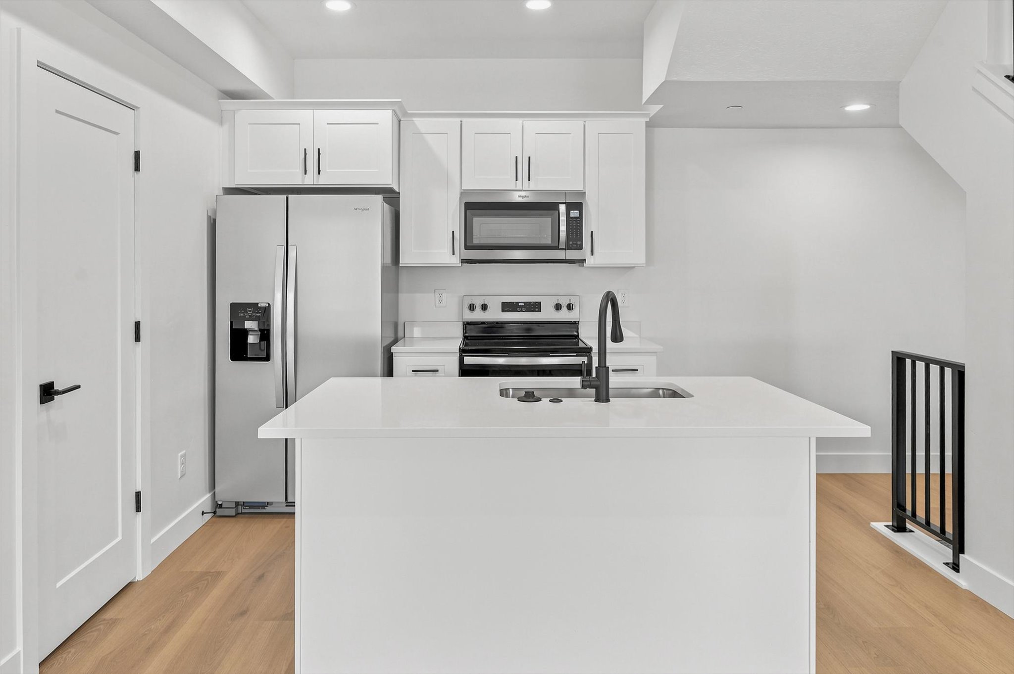 Modern kitchen featuring an island with an integrated sink and gooseneck faucet, light wood-finish flooring, white cabinetry, stainless steel appliances, and recessed lighting