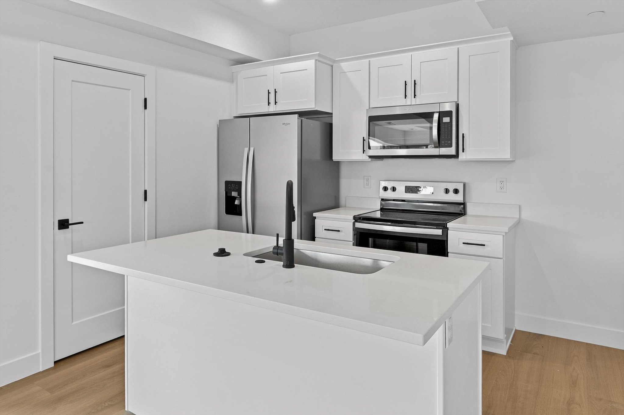 Kitchen featuring a center island with an undermount sink and matte black faucet, white shaker cabinetry, stainless steel appliances, and light wood-finish flooring