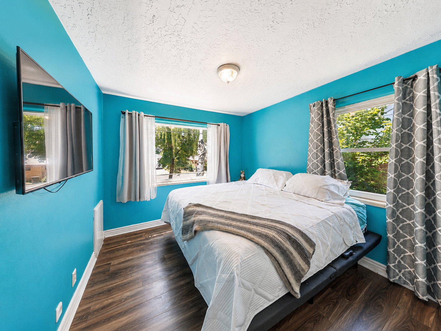 Bedroom featuring wood-finish flooring, two windows with window treatments, a wall-mounted display, and baseboard trim