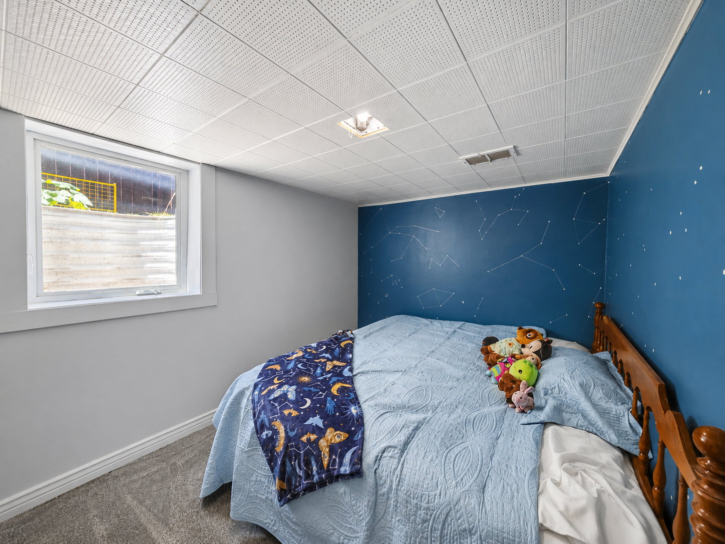 Room featuring a drop ceiling with recessed lighting, blue accent wall with constellation motif, gray carpeting, and a white-framed window