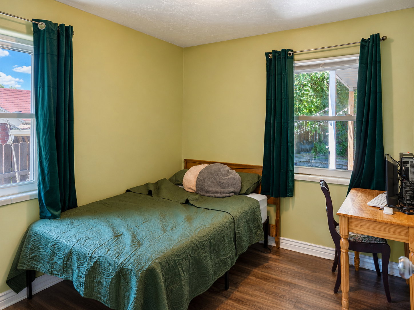 Bedroom featuring wood-finish flooring, two windows with deep green drapes, light yellow wall color, and white baseboards